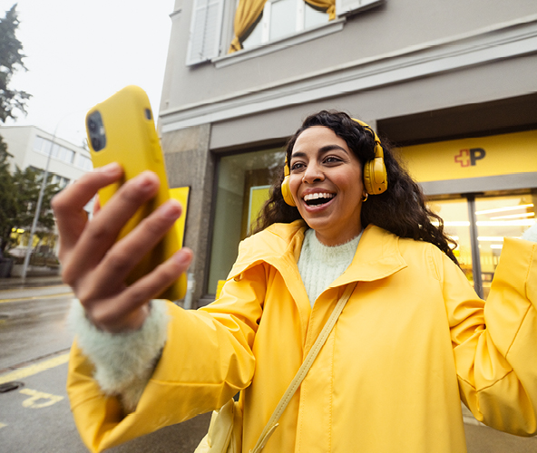 Young woman staring in front of a Post Mobile office, in a videocall with the family abroad. She's using a Post Mobile subscription. 